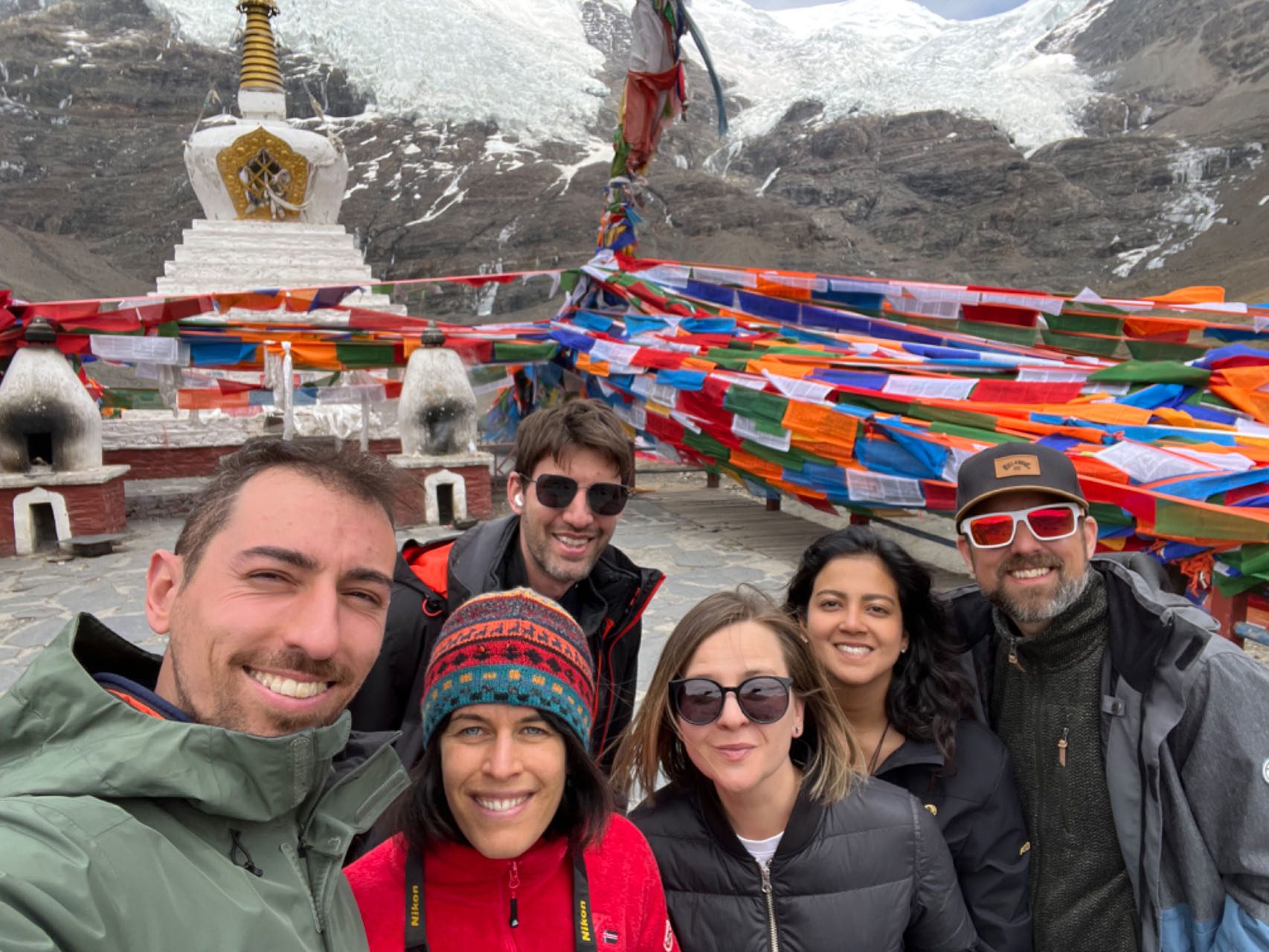 Solo Travelers at Sacred Stupa with Prayer Flags - Tibet Spiritual Solo travelers group smiling at sacred Buddhist stupa with colorful prayer flags and Himalayan glacier background