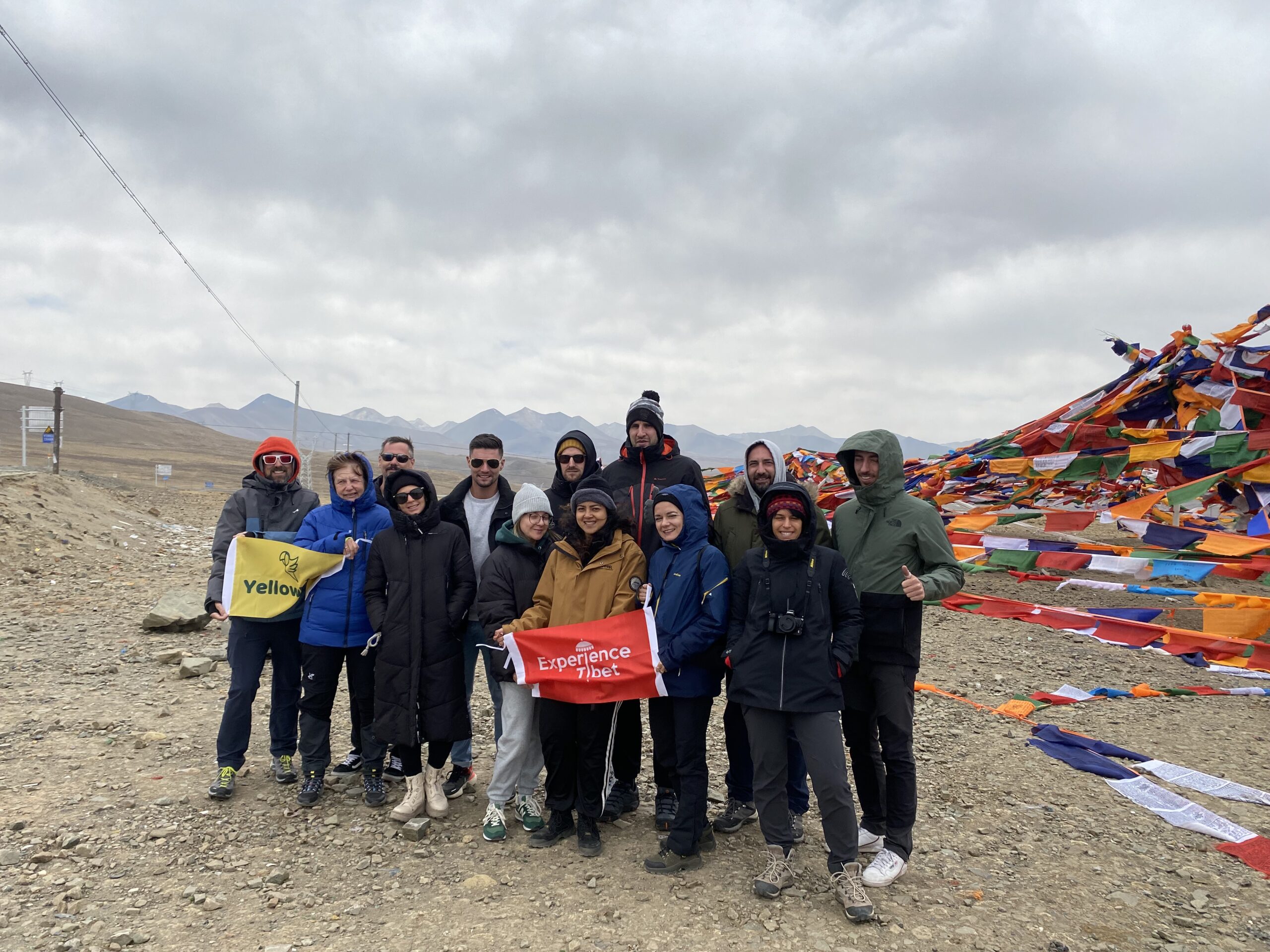 Solo Travelers with Experience Tibet Flag - High Altitude Community Solo travelers group holding Experience Tibet and YellowBird Tour flags at high altitude plateau with colorful prayer flags background