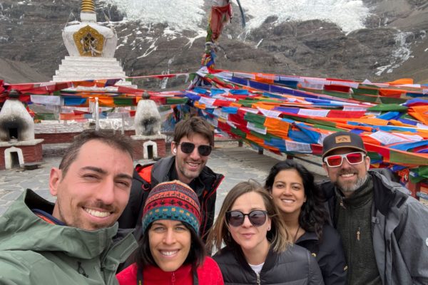 Solo Travelers at Sacred Stupa with Prayer Flags – Tibet Spiritual Solo travelers group smiling at sacred Buddhist stupa with colorful prayer flags and Himalayan glacier background